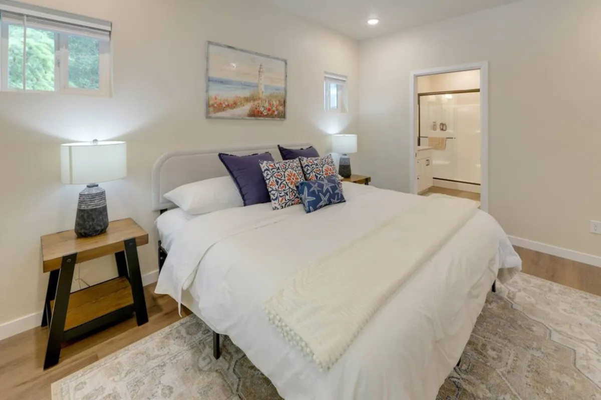 A serene, sun-lit bedroom featuring a queen bed adorned with decorative pillows, landscape artwork above the headboard, and matching bedside lamps. Sunlight streams through a window, creating a cozy atmosphere ideal for relaxation and restful sleep.