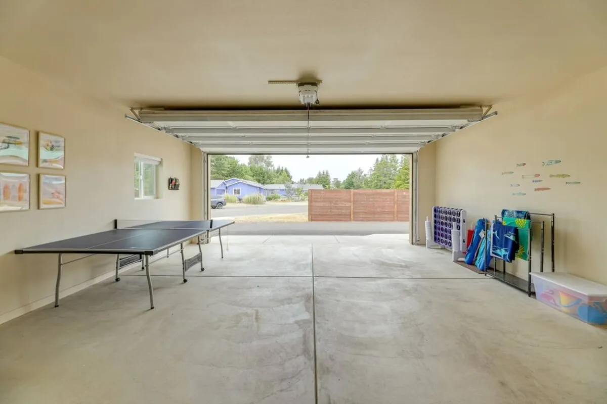 A bright game room featuring a ping pong table as the centerpiece, with a garage door in the background, storage racks, and colorful beach items lining the walls. The space exudes a playful, casual vibe and is clearly designed for active fun and family gatherings.