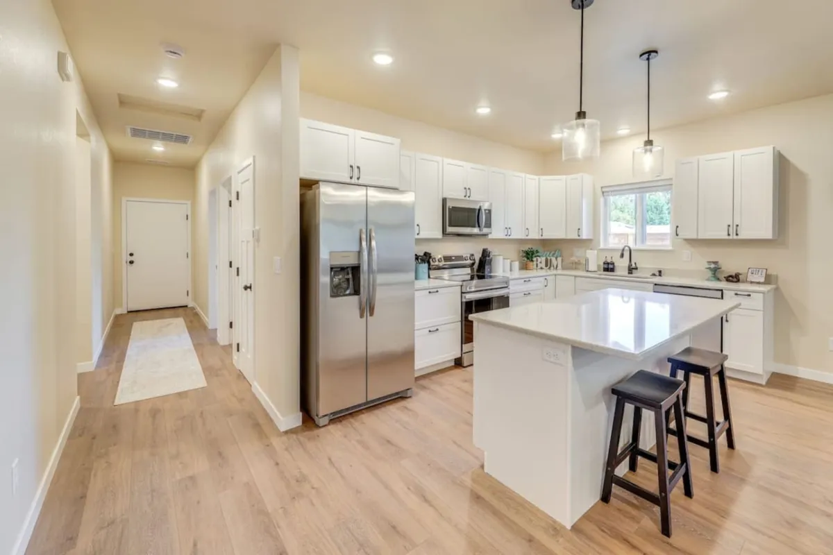 A bright, modern kitchen featuring a spacious island, gleaming stainless steel appliances, and crisp white cabinetry. The scene feels warm and welcoming, with hints of cookware visible, inviting gatherings and culinary creativity.