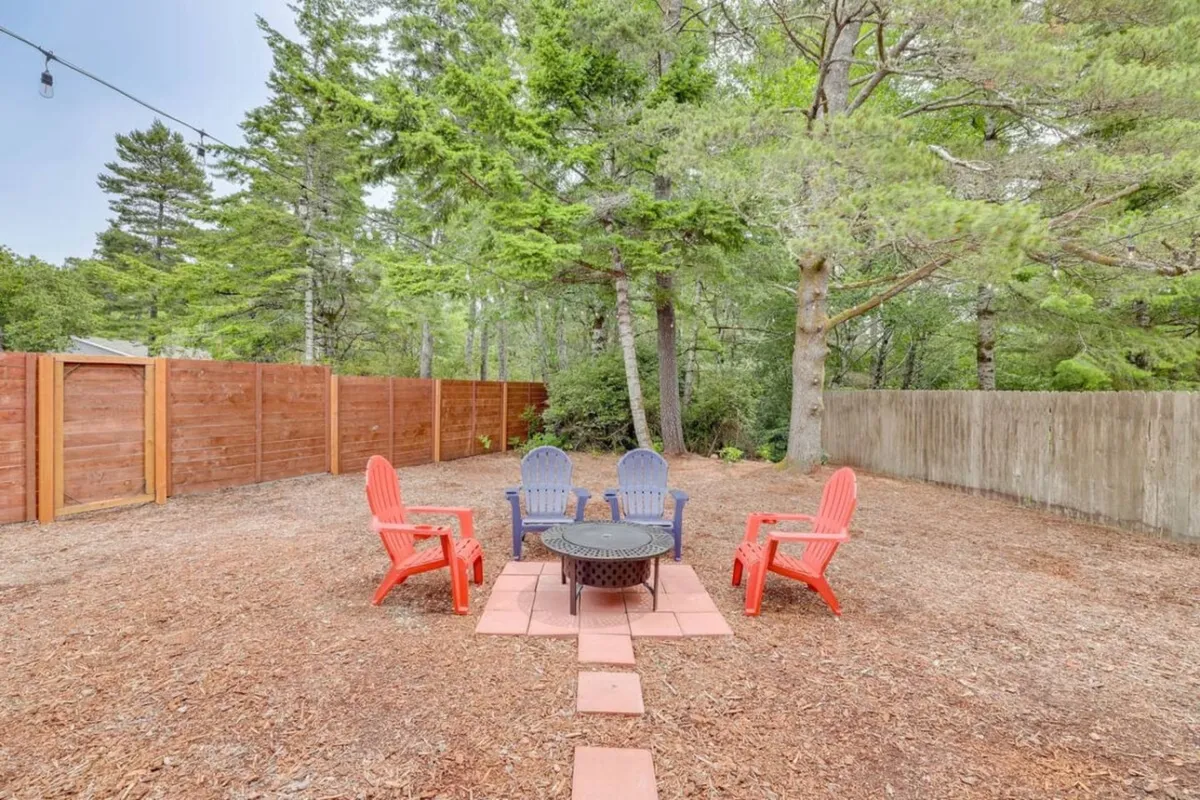 The image features the cozy outdoor space of Duck Creek Cottage, showcasing a fire pit surrounded by inviting red and blue chairs on a rustic patio, all framed by a wooden fence and towering trees. The setting is perfect for evening gatherings, relaxation, and connecting with friends or family under the stars.