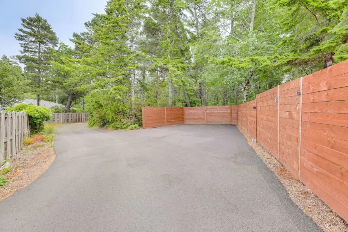 This exterior image captures a paved driveway surrounded by lush trees and a wooden fence. The natural setting hints at the tranquil, forested environment that characterizes the area and provides space for outdoor activities and play.