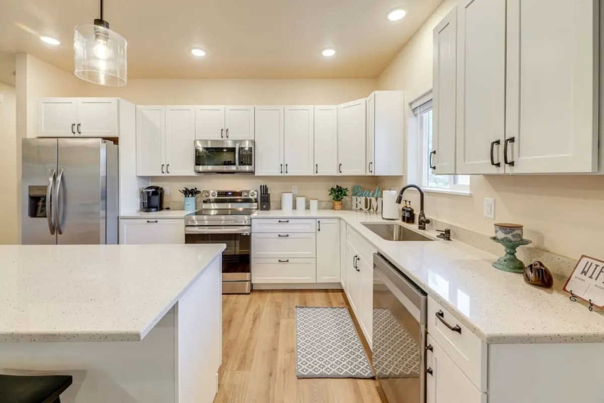 A wide view of the kitchen area with a prominent island, stainless steel appliances, white cabinetry, and a sink beneath a sunny window. The open layout hints at easy flow to adjoining spaces, ready for both cooking and entertaining.