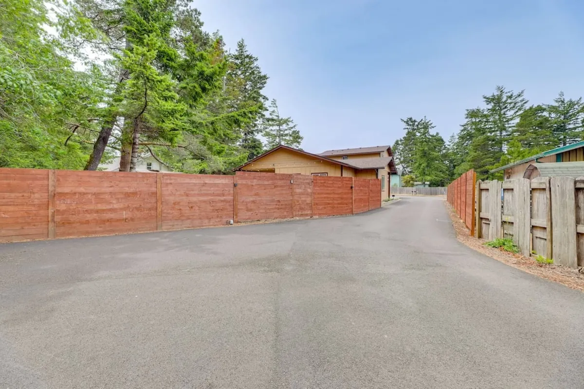 This exterior scene shows a wooden fence bordering a leafy area, with a roadway leading toward the house. The view through the trees provides a feeling of peaceful seclusion and connection to nature trails in the surrounding neighborhood.